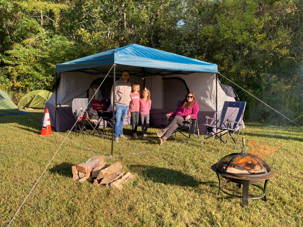 Family posing under a blue camping canopy with a tent, chairs, and a campfire nearby in a grassy field.