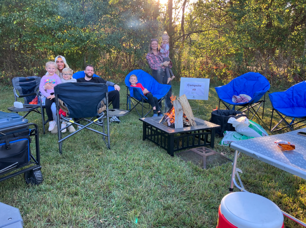 Family group sits around a small campfire on a grassy field with blue and black camping chairs and a sign reading Campsite #9, enjoying an outdoor evening.
