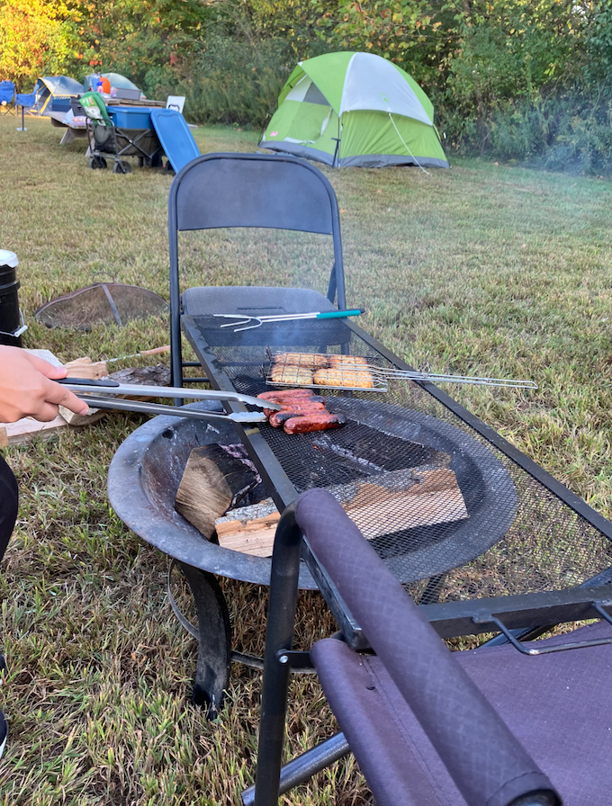 Campground barbecue scene with a green tent in the background; meat cooks on a metal mesh grill over a wood-burning fire pit, tongs in use.