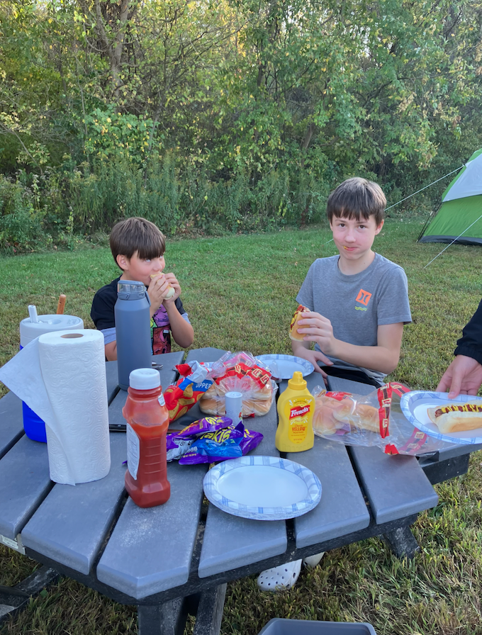 Two boys sit at a picnic table outdoors during a camping trip, eating hot dogs with chips and condiments nearby while a tent is visible in the background.
