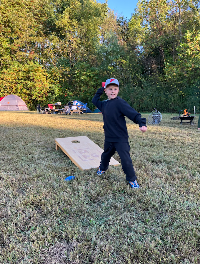 Young boy in a black outfit throws a bean bag toward a cornhole board at a family campsite. Autumn trees and tents in the background.