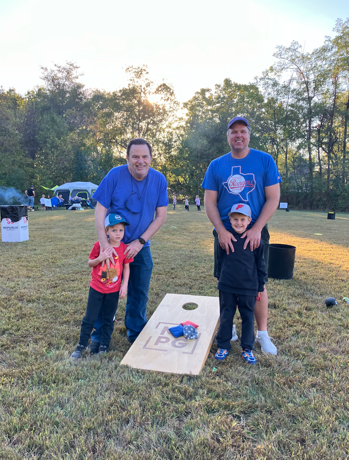 Two men with two young children posing beside a cornhole board on a sunny outdoor field with tents in the background.