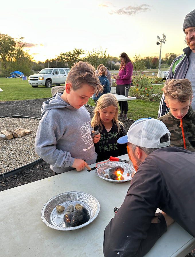 Children gather around a picnic table outdoors as a man with a cap tends to a small campfire in foil pans during a park camp activity at sunset, with a girl and two boys watching closely.