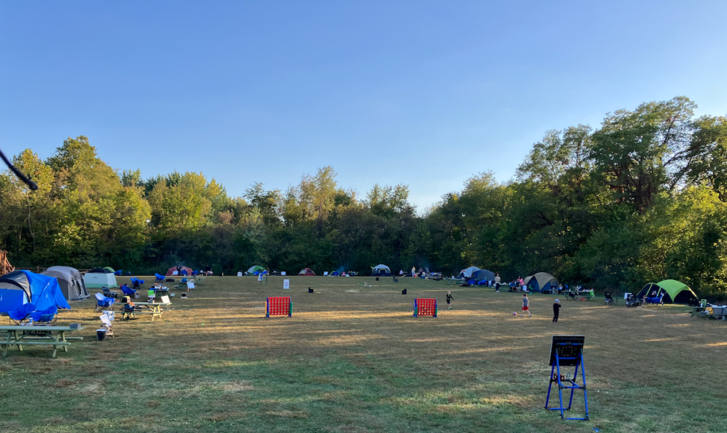 Open campground field with numerous tents along a tree line and people walking; two red Connect Four boards in the center.